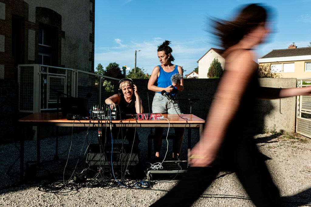 Une photo du spectacle. une femme passe au premier plan elle est floue. Au second plan deux femmes derrière une table avec du matériel radiophonique. 